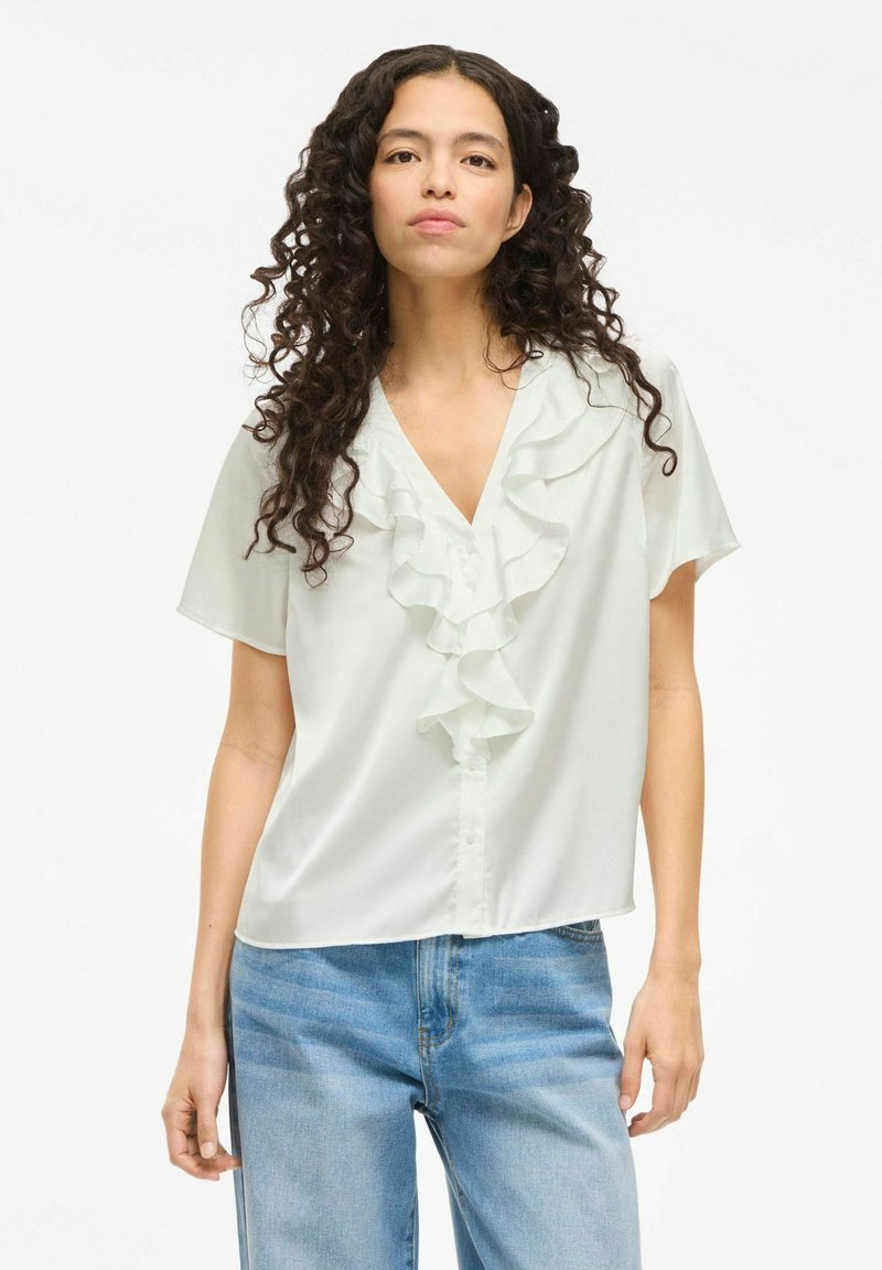 Hvid kortærmet bluse med V-hals og faldende flæser ned ad foran. Stoffet ser glat og let blankt ud. Casual pasform.