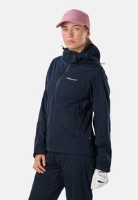 Femme portant une veste Swedemount bleu marine, un pantalon assorti, un gant de golf blanc Wilson et une casquette de baseball rose, debout devant un fond uni.