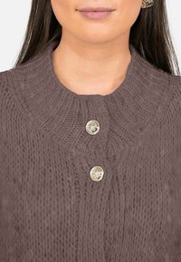 Pull en tricot marron à col haut, avec des motifs texturés et deux boutons métalliques au niveau du col. Matière douce et épaisse.