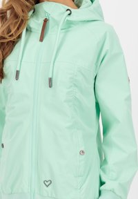 Mintgroene lichte jas met een rits aan de voorkant, een capuchon en verstelbare trekkoorden. Heeft zijzakken en een subtiel hartdetail.