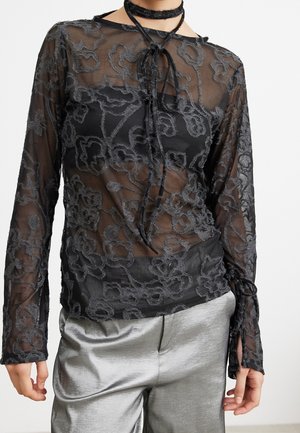 Frau trägt ein schwarzes, transparentes, langärmeliges Oberteil mit floraler Stickerei, darunter ein schwarzes Trägertop und silberne Satinhose.