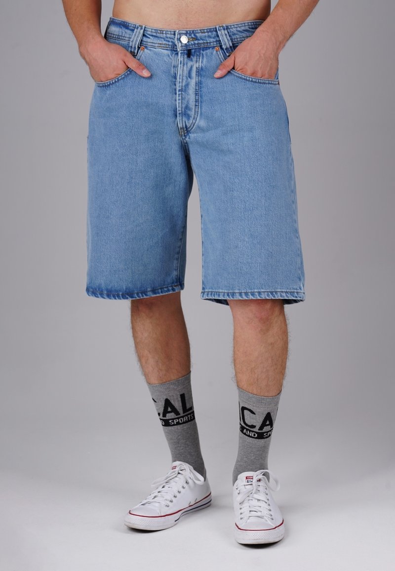 Light blue denim shorts with a relaxed fit, featuring a button closure, two side pockets, and a hemmed edge. Paired with gray socks and white sneakers.