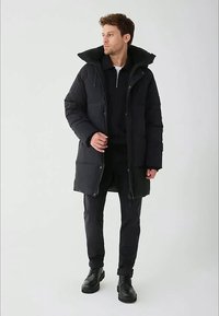 Mężczyzna w czarnej puchowej kurtce, czarnych spodniach i czarnych butach, stoi i patrzy w bok na tle jednolitego szarego tła.