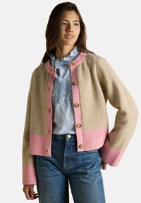 Žena v béžovom kardigáne s ružovým lemovaním a gombíkmi, oblečená v modrej blúzke s volánmi, v kombinácii s modrými džínsami, stojí s jednou rukou na bok.