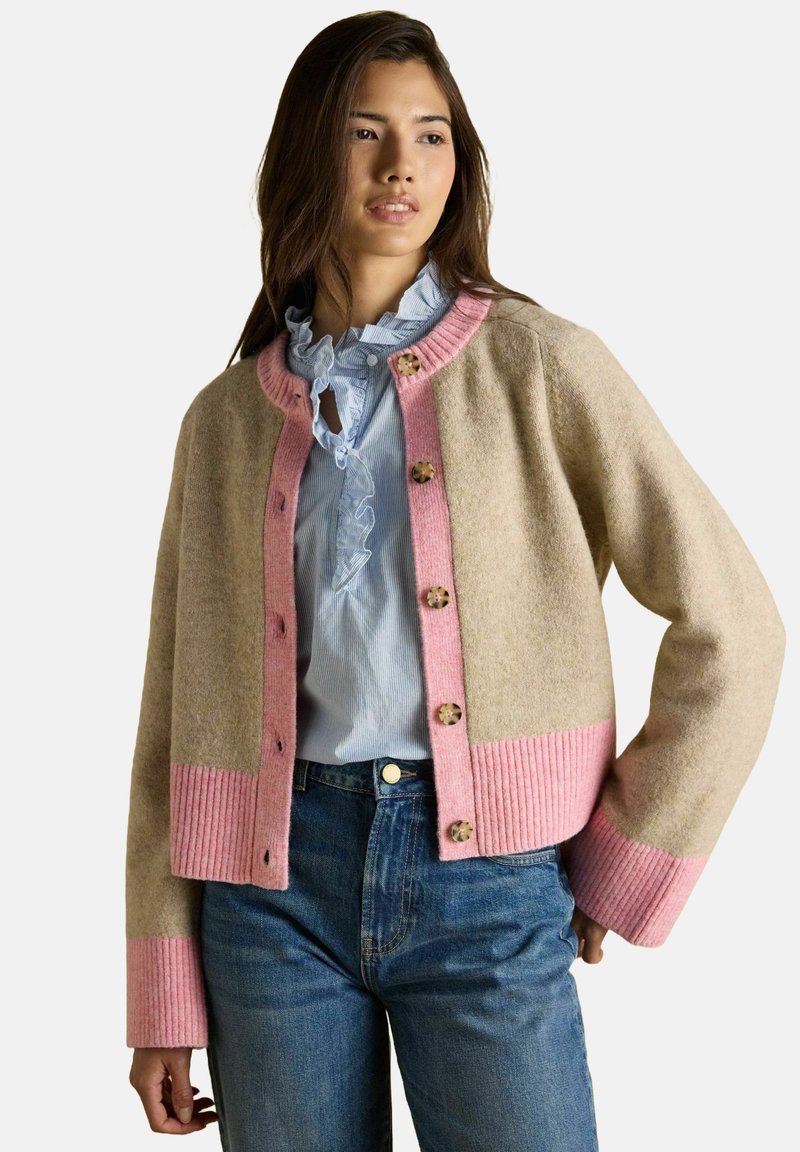 Žena v béžovom kardigáne s ružovým lemovaním a gombíkmi, oblečená v modrej blúzke s volánmi, v kombinácii s modrými džínsami, stojí s jednou rukou na bok.