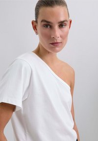 Weißes Baumwoll-T-Shirt mit asymmetrischem Ausschnitt, kurzen Ärmeln und weicher Textur. Das Design hat eine Schulter frei.