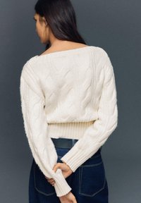 Fehér kötött pulóver laza vállvonallal és bordázott mandzsettákkal, sötét magasderekú farmernadrággal párosítva.