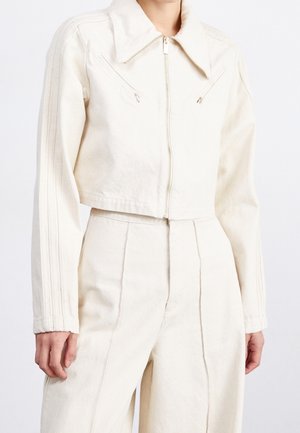 Mujer con chaqueta corta color blanco roto con cremallera frontal y pantalones de talle alto a juego sobre un fondo blanco liso.