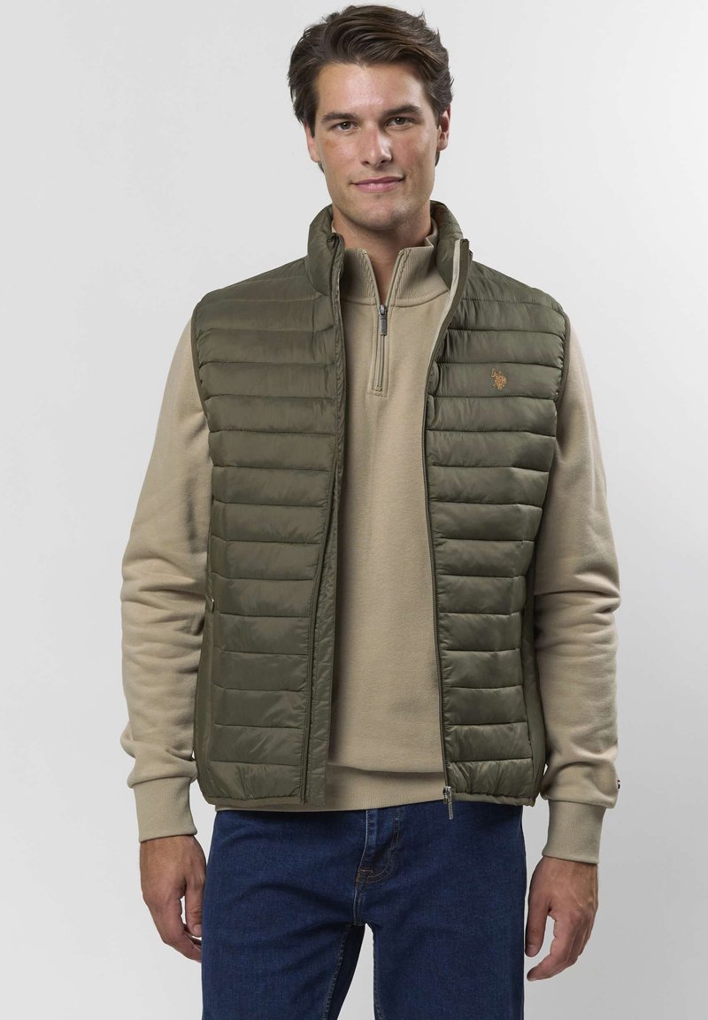 Mężczyzna ubrany w oliwkowozieloną pikowaną kamizelkę na beżowym swetrze z półgolfem i niebieskich dżinsach, stojący na jednolitym tle.