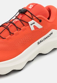 Heldere oranje sportieve schoen met een bovenwerk van mesh, zwarte veters en een dikke witte zool met een gestructureerd patroon. "SALOMON" logo aan de zijkant.