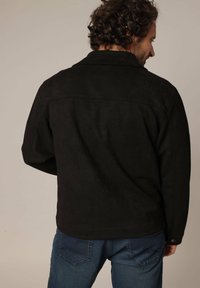 Veste en daim noir au fit décontracté, avec une exposition de coutures et des détails de boutons sur les manches ; vue de dos.