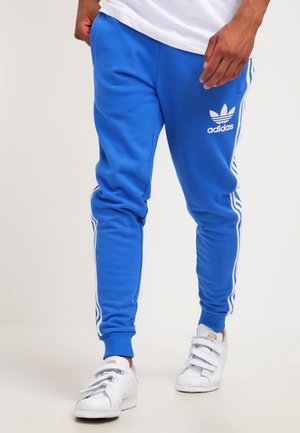 Mężczyzna ubrany w niebieskie spodnie dresowe Adidas z białymi paskami i białe trampki, z rękami w kieszeniach, stojący na jednolitym tle.