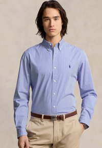 Camisa de botones a rayas en azul y blanco, con un cuello clásico, mangas largas y un pequeño logo en el pecho; combinada con pantalones beige.