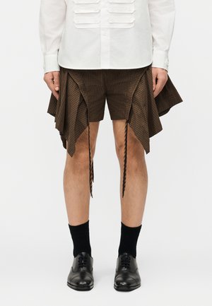 Homme portant un short marron à carreaux asymétriques avec des panneaux drapés, des chaussettes noires, des chaussures habillées noires et une chemise blanche à manches longues boutonnée.