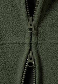 Olivengrønn fleecejakke med et foret, teksturert overflate og to grå glidelåser foran, med diskré sømmer.