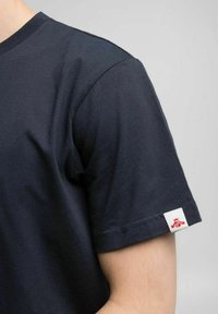 T-shirt in cotone blu navy con collo rotondo, maniche corte e un'etichetta bianca con un logo rosso sulla manica sinistra. Tessuto liscio.