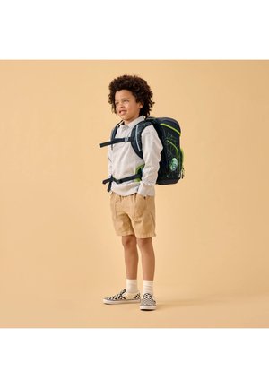 Child with curly hair wearing a gray sweatshirt, tan shorts, checkered slip-on shoes, and a black and green backpack standing on a beige background.