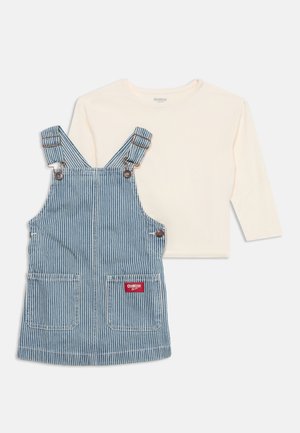 Chemise crème à manches longues associée à une robe tablier rayée bleue et blanche avec poches avant et bretelles réglables à boucle.