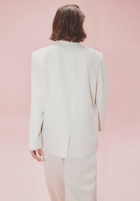 Blazer cintré beige clair avec des épaules structurées, une fente au dos et une texture lisse, associé à un pantalon assorti sur fond rose.