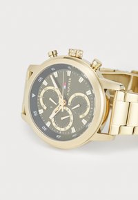 Montre-bracelet dorée avec un cadran noir, trois sous-cadrans, affichage de la date et bracelet en maillons métalliques, portant le logo Hilfiger.