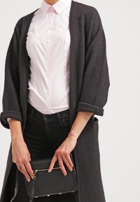 Cardigan nero in misto lana con maniche arrotolate, indossato sopra una camicia rosa chiaro con bottoni e jeans neri, mentre tiene una piccola borsa nera.