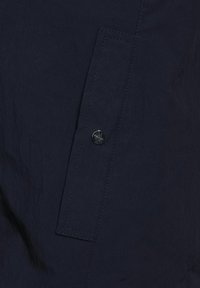 Mörkblått tyg med en slät yta som har en knapp med en logotyp, sydd fickdetalj och synliga sömmar.