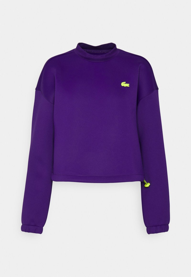 Sweat-shirt violet coupé court avec des manches longues, un col haut et un logo vert sur la poitrine. Comprend des poignets élastiques et une texture lisse.