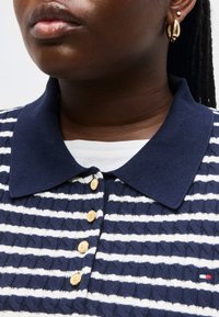 Κοντινό πλάνο ενός ατόμου που φοράει πλεκτό πόλο με ναυτικό μπλε και λευκές ρίγες, με χρυσά κουμπιά, μικρό λογότυπο και χρυσά κρίκους και σκουλαρίκια καρφίτσα.
