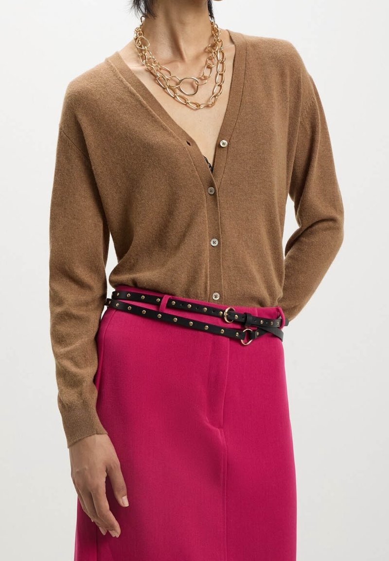 Femme portant un cardigan marron boutonné rentré dans un pantalon rose vif avec double ceinture noire cloutée et un collier chaîne épais en or.