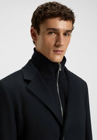 Schwarzer Wollmantel mit hohem Kragen und Reißverschluss. Glatte Textur und taillierte Passform, mit minimalistischem Design.