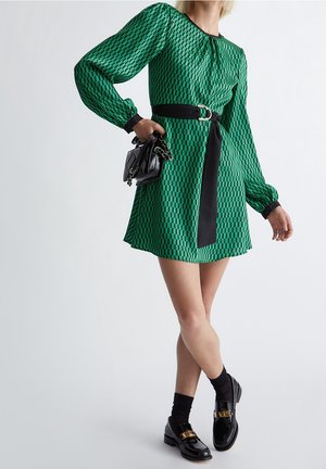 Femme portant une robe verte à motif avec des manches longues et une ceinture noire, tenant un sac à main noir, portant des chaussettes noires et des mocassins brillants.