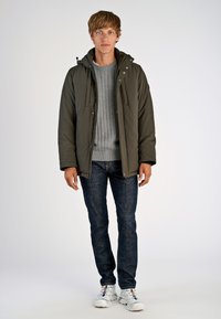 Pufferjakke i mørkegrøn, med hætte, lynlås-lommer og en tekstureret grå sweater under, parret med mørke jeans og hvide sneakers.