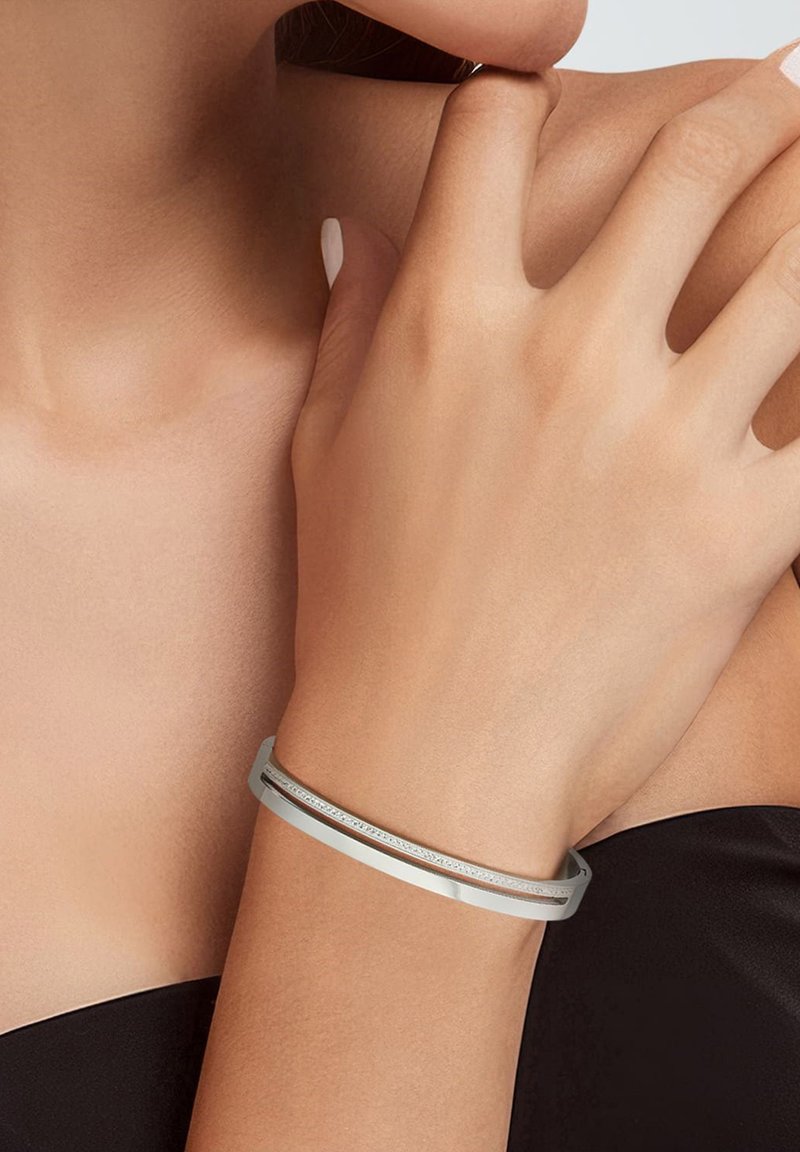Pulsera de bangle de plata con un acabado suave y un diseño sutil, llevada en una mano con color de uñas neutro contra un top oscuro.