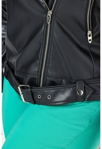 Zwarte leren jack met zilveren ritsen, aansluitend ontwerp en een brede riem met een gesp. Draagt over fel teal-kleurige kleding.