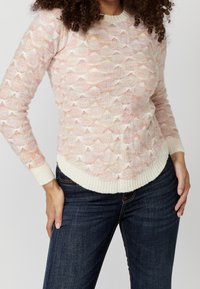 Suéter de punto en rosa pálido, con un patrón de olas texturizado, cuello y puños acanalados en crema, y un dobladillo ligeramente curvado. Combinado con jeans oscuros.