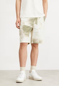 Light-colored shorts made from textured fabric with a grid pattern in green. Features side pockets and a relaxed fit. Worn with white sneakers.