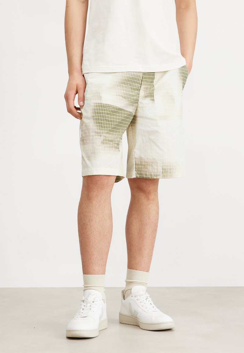 Light-colored shorts made from textured fabric with a grid pattern in green. Features side pockets and a relaxed fit. Worn with white sneakers.
