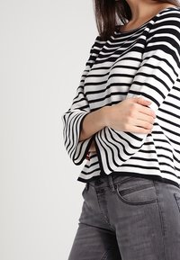 Haut à manches longues rayé noir et blanc avec manches évasées, associé à un jean gris. Le tissu semble doux avec une silhouette ajustée.