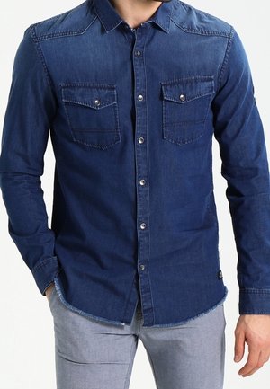 Mężczyzna ubrany w ciemnoniebieską denimową koszulę na guziki z dwoma przednimi kieszeniami na piersi oraz jasnoszare spodnie, ręce swobodnie opuszczone wzdłuż ciała.