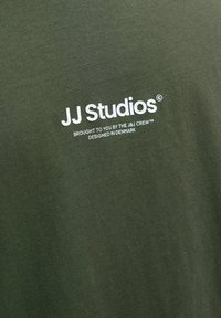 Olíva zöld, bordázott textúrájú póló, fehér nyomattal: "JJ Studios" és szlogen. Egyszerű dizájn, rövid ujjú.