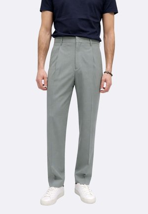 Uomo che indossa pantaloni sartoriali verde salvia, sneakers bianche e una maglietta blu scuro, in piedi con le braccia rilassate ai lati.