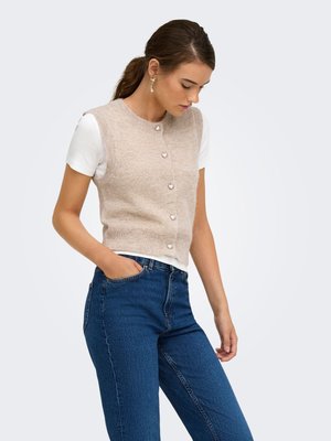 Femme portant un gilet tricoté beige sans manches avec des boutons en forme de cœur par-dessus un t-shirt blanc et un jean bleu, regardant vers le bas avec une main dans la poche.