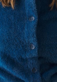 Niebieski, puszysty kardigan o miękkiej teksturze, z czterema dopasowanymi, okrągłymi guzikami z przodu. Zbliżenie podkreśla szczegóły guzików.