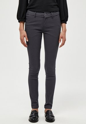 Femme portant un pantalon slim gris foncé, un haut noir à manches longues et des mocassins noirs, debout devant un fond clair uni.