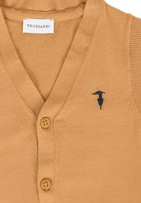 Beżowy, dzianinowy kardigan bez rękawów z dekoltem w serek, dwoma guzikami i małym, ciemnym haftowanym logo na klatce piersiowej.