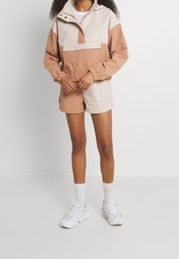 Kevyt, beige ja persikanvärinen anorakki, jossa puolivetoketju, joustavat hihansuut ja yhteen sopivat shortsit. Yhdistetty valkoisiin tennareihin ja sukkiin.