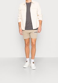 Beige šortky s logem, šedé tričko a krémová kordurová košile. Doplňuje bílé tenisky a ponožky, čímž vytváří ležérní outfit.