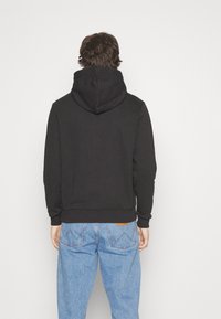 Black hoodie with a kangaroo pocket and adjustable drawstring hood. Paired with light blue denim jeans featuring a leather patch.