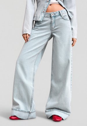 Osoba nosiaca svetlomodré rifle s širokými nohavicami, zodpovedajúcu skrátenú riflovú bundu, biely top a červené špicaté topánky na jednoduchom pozadí.