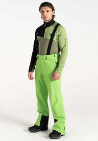 Ski-Outfit mit einem schwarzen und olivgrünen Oberteil mit Stehkragen, kombiniert mit leuchtend grünen wasserfesten Hosen und schwarzen Skischuhen.